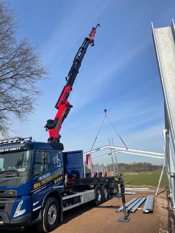 Blauwe kraanwagen van Constructie De Greyt met rode Palfinger-kraan die stalen spanten optilt
